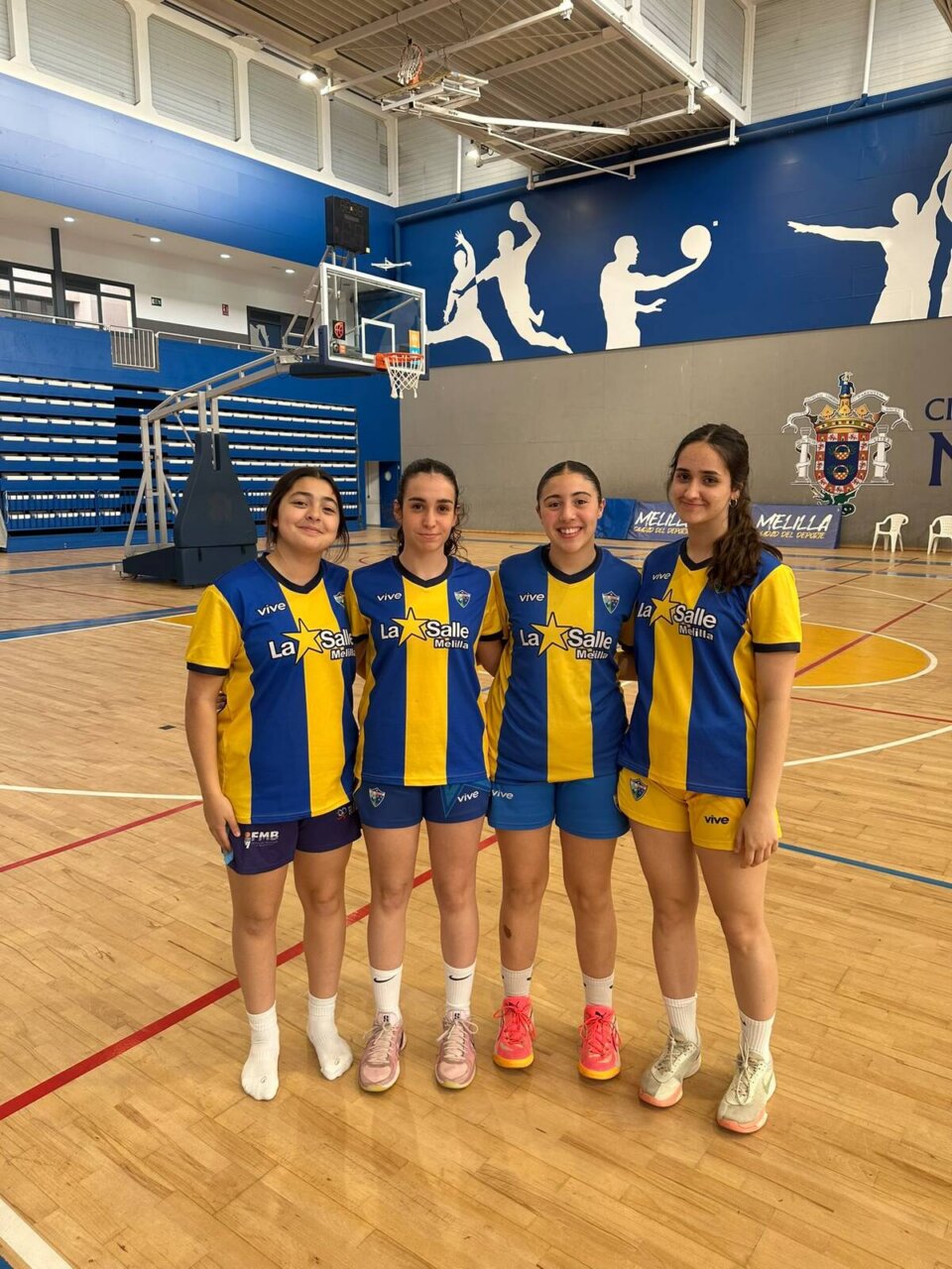 Four young female athletes in blue and yellow La Salle Melilla uniforms pose together on an indoor basketball court, smiling for a team photo.