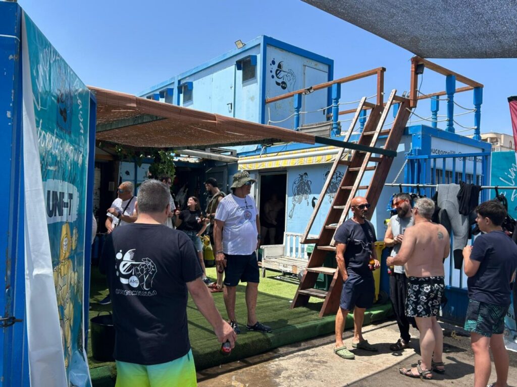 Group of people standing and chatting in a sunny outdoor area with blue shipping-container buildings and a wooden staircase.