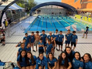Group of young swimmers and coaches in matching blue shirts posing on a pool deck in front of a large outdoor swimming pool with blue lane lines and a curved translucent roof structure behind them.