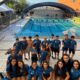Group of young swimmers and coaches in matching blue shirts posing on a pool deck in front of a large outdoor swimming pool with blue lane lines and a curved translucent roof structure behind them.