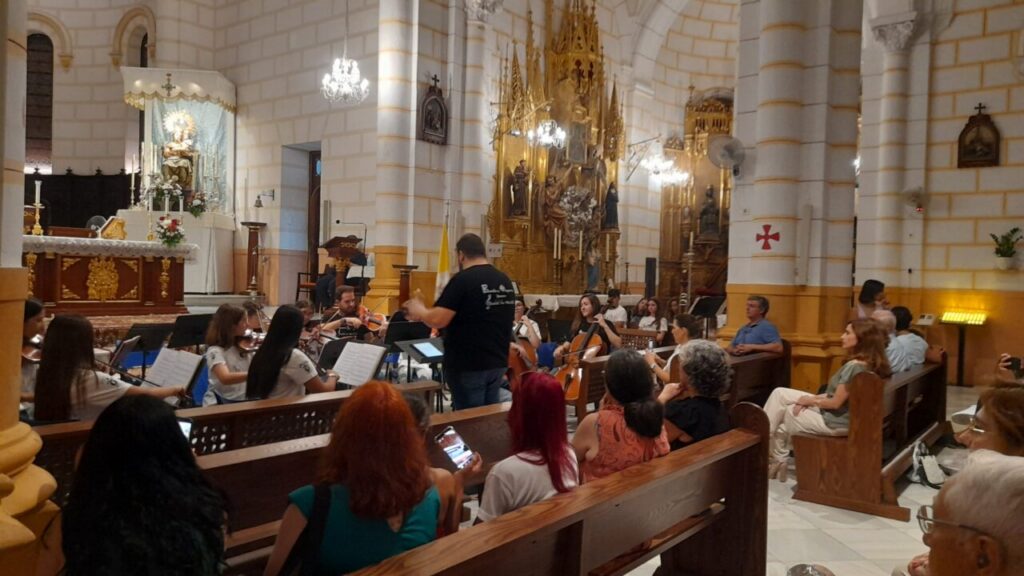 String orchestra performing in a church nave, conductor directs players as audience watches from wooden pews, chandeliers above.
