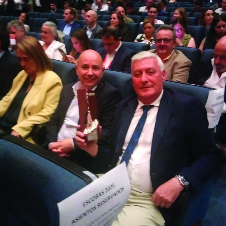 Two men in business attire sit in an auditorium; one bald man holds a shiny trophy while the other wears a blue tie. Behind them, an audience watches the event.