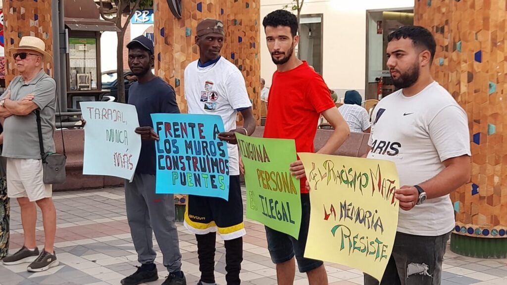 Group of five men standing in a plaza, each holding colorful protest signs with messages in Spanish (anti-racism/immigration). The blue sign reads: 'Frente a los muros construimos puentes'; the green sign says 'Ninguna persona ilegal'; the yellow sign says 'El racismo mata la memoria, resiste' (partial reads visible); a man in red stands near the center.