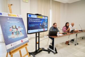Panel discussion at a cybersecurity workshop: three speakers sit at a table with laptops and microphones, a presentation screen behind them, and a large poster on an easel to the left.