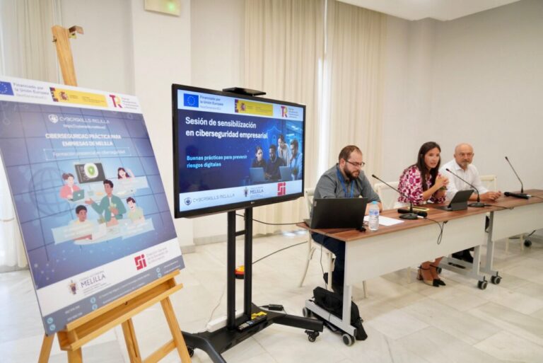 Panel discussion at a cybersecurity workshop: three speakers sit at a table with laptops and microphones, a presentation screen behind them, and a large poster on an easel to the left.