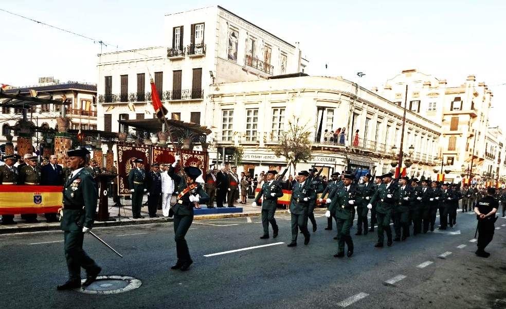 Military parade on a city street with uniformed soldiers marching, banners and spectators along the curb.