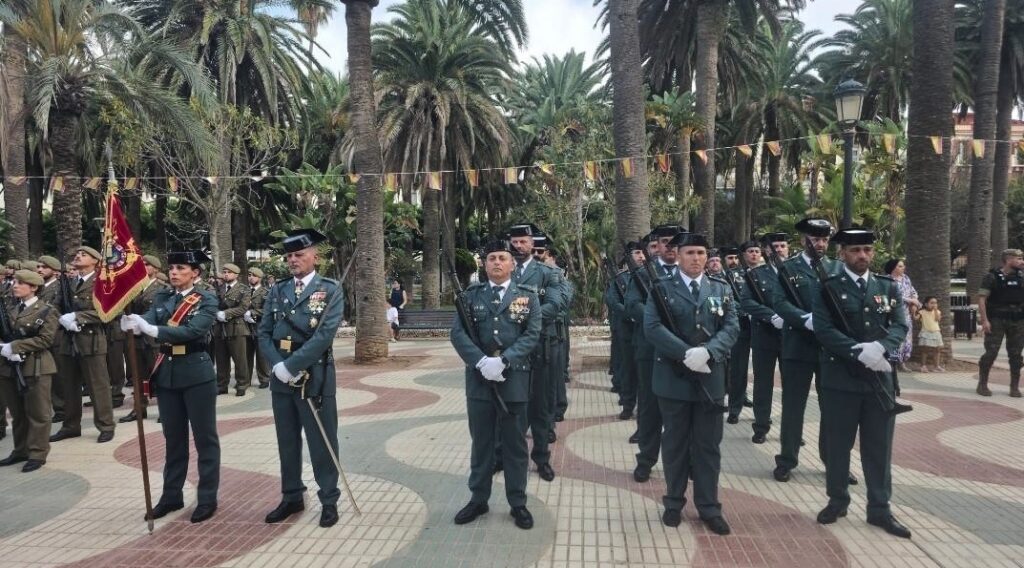 Group of soldiers in green dress uniforms standing at attention in a palm-lined plaza during a ceremony, with a flag bearer on the left and spectators in the background.