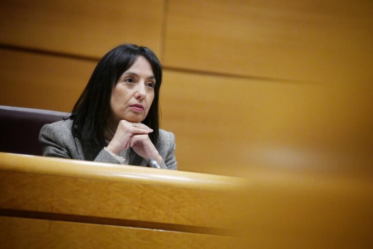 Woman with dark hair in a gray blazer sits at a wooden bench, hands clasped under chin, looking thoughtful in a formal setting.