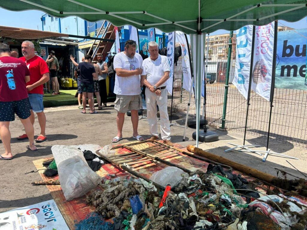 Two men under a green canopy discuss near a pile of sea-waste and debris laid out on a striped mat at an outdoor event.