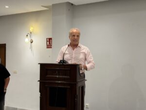 Man in a pink shirt speaks at a dark wooden podium with a microphone in a conference room.