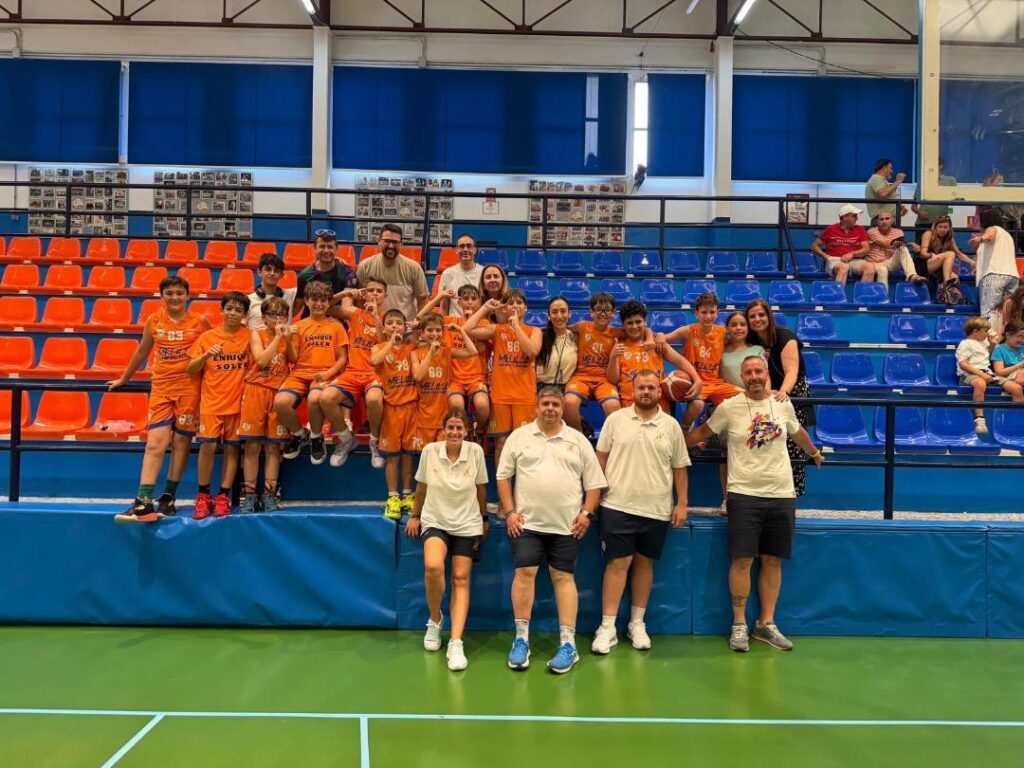 Youth basketball team in orange uniforms posing for a group photo on blue bleachers inside a gymnasium, with coaches standing behind them and a few spectators in the background.