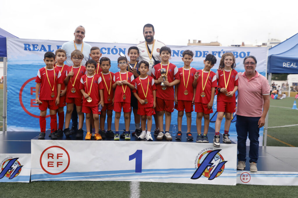 Youth soccer team on a podium wearing red uniforms with medals, posing with coaches and a trophy after a win event.