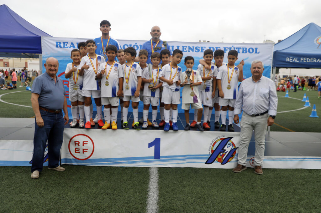 Group of young soccer players in white uniforms with gold medals on a first-place podium, flanked by two men, at a field with banners in the background.