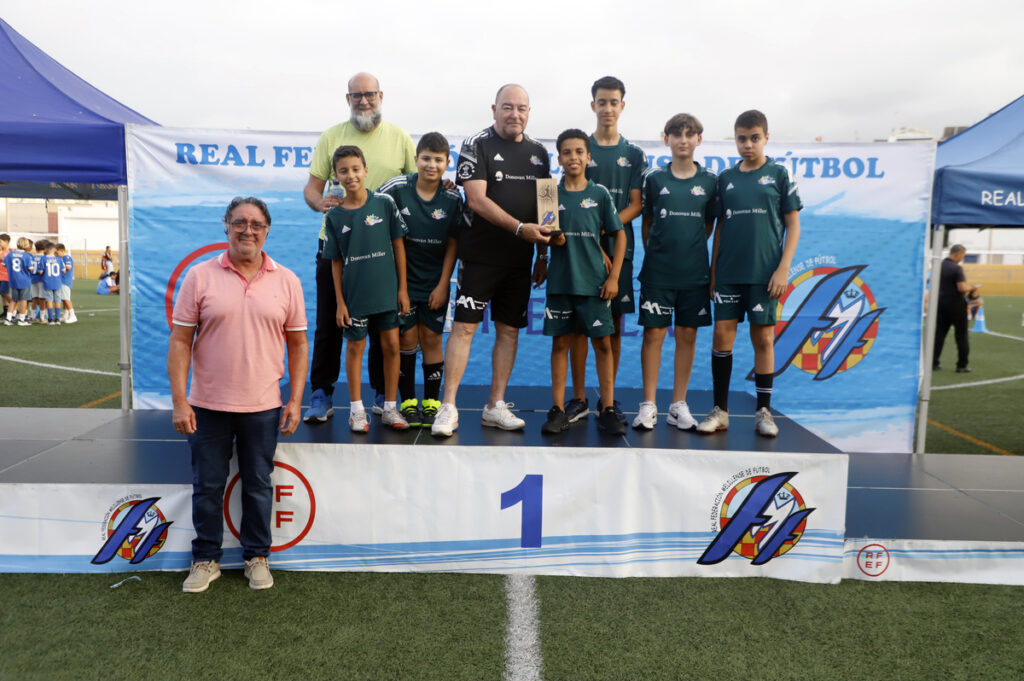 Group of young soccer players in green uniforms on a winner's podium with a trophy, flanked by two adults, on a football field with banners in the background.