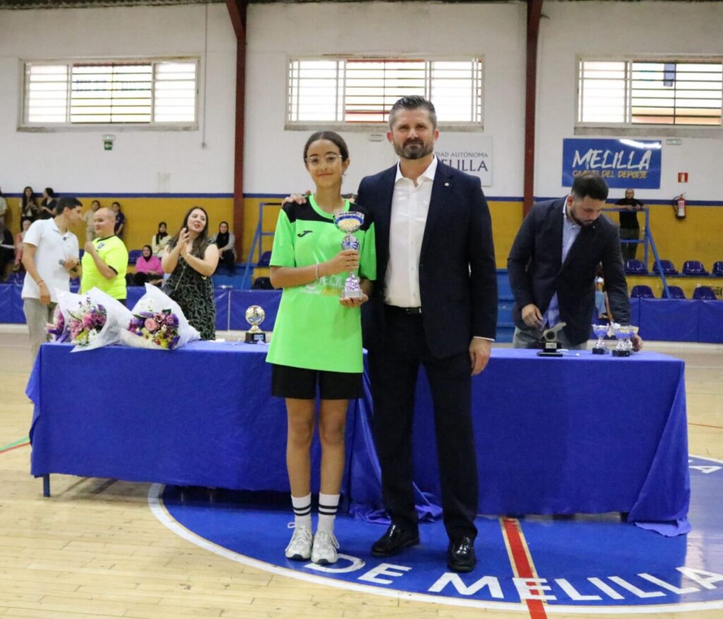 Girl in a bright green sports jersey holds a trophy beside a suited man at an indoor awards ceremony on a gym floor.