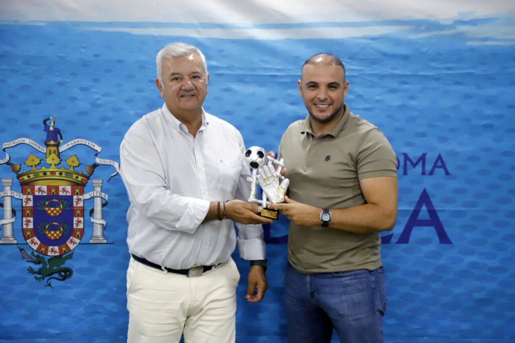 Two men present a soccer trophy in front of a blue backdrop with a coat of arms in the background, handshaking as they hold the award.