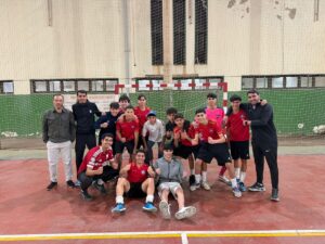 Group of youth soccer players in red jerseys posing with two coaches on an indoor court in front of a goal net.
