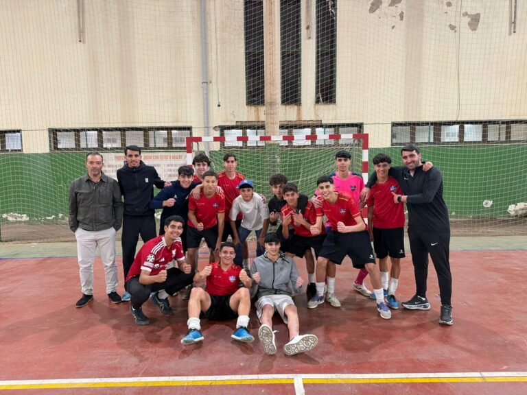 Group of youth soccer players in red jerseys posing with two coaches on an indoor court in front of a goal net.