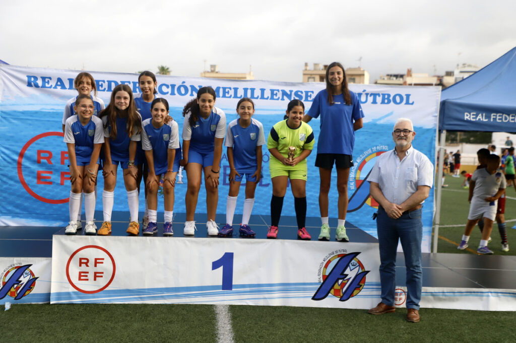 Youth girls' soccer team wearing blue and white uniforms pose on a first-place podium with a trophy, joined by a smiling man in a light blue shirt on a sports field backdrop.