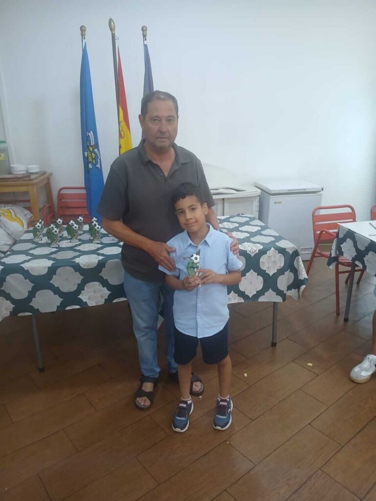 Man stands with a boy who is holding a small trophy in a room set up for awards, with flags and tables in the background.”,