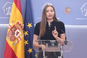 Speaker at a podium gesturing with one finger, flanked by the Spanish and EU flags in a congressional press room (Congreso de los Diputados).