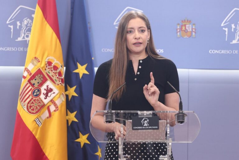 Speaker at a podium gesturing with one finger, flanked by the Spanish and EU flags in a congressional press room (Congreso de los Diputados).