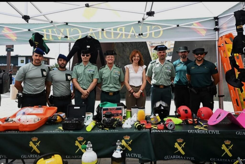 Guardia Civil officers plus a civilian woman posing behind a table of safety gear under a canopy; rescue equipment and helmets displayed in front of them.