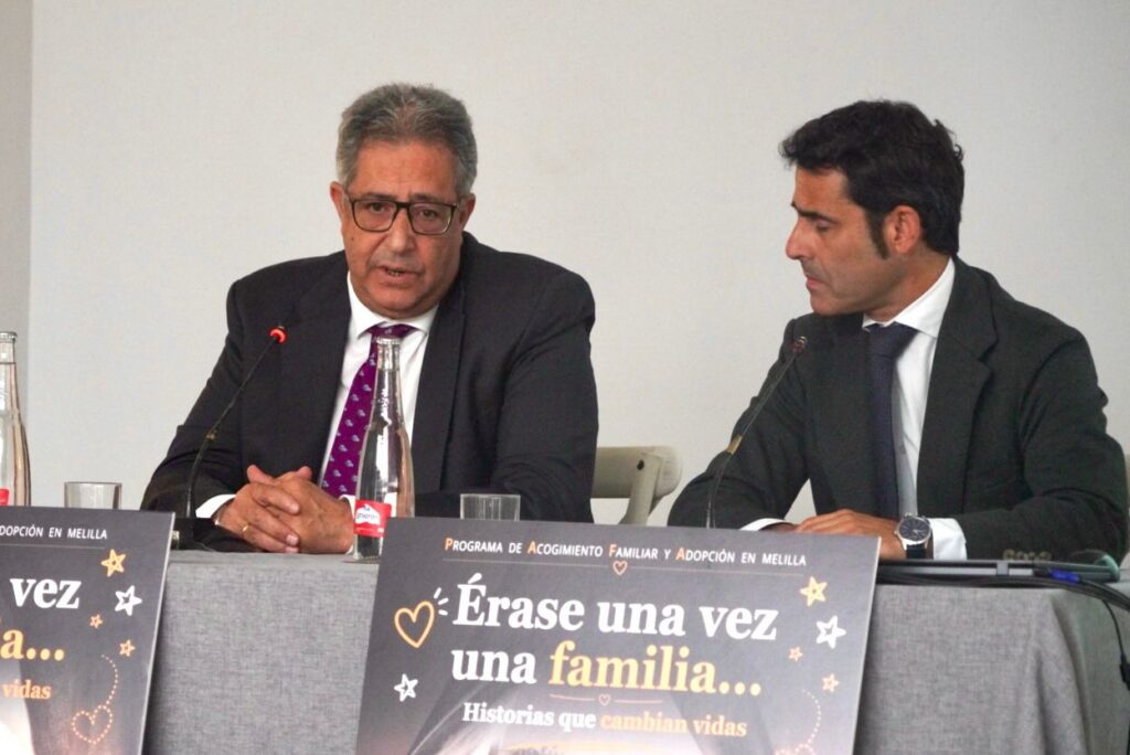 Two men in suits sit at a panel table speaking into microphones, with a glossy poster in the foreground reading 'Érase una vez una familia... Historias que cambian vidas'.