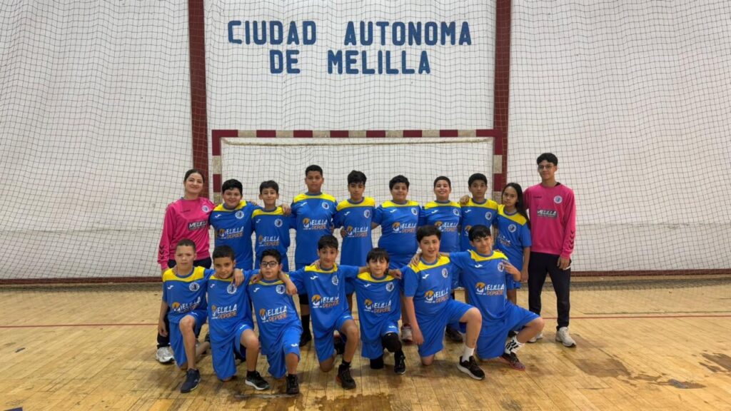 Group of young futsal players in blue and yellow uniforms posing with two coaches in pink jerseys in a gym, goal and net behind them, banner reads Ciudad Autónoma de Melilla.