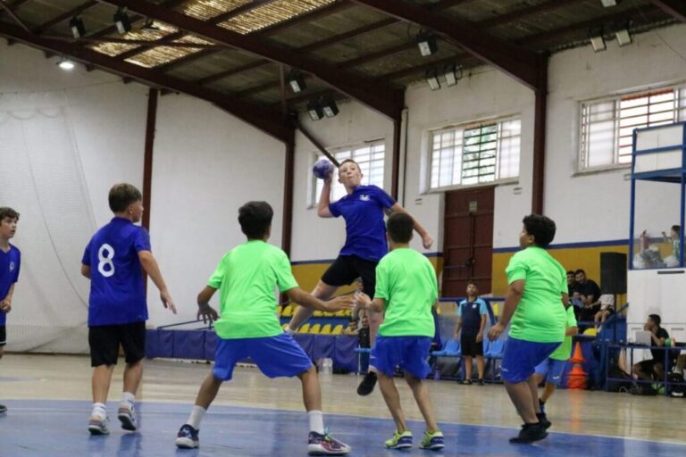 Youth indoor team sport: a blue-shirted player leaps to throw a ball while others in green shirts surround him. A gymnasium with spectators sits in the background.