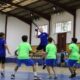 Youth indoor team sport: a blue-shirted player leaps to throw a ball while others in green shirts surround him. A gymnasium with spectators sits in the background.