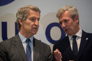 Two men in dark suits and ties converse on a stage, a blue-and-white backdrop behind them highlighting partial letters.