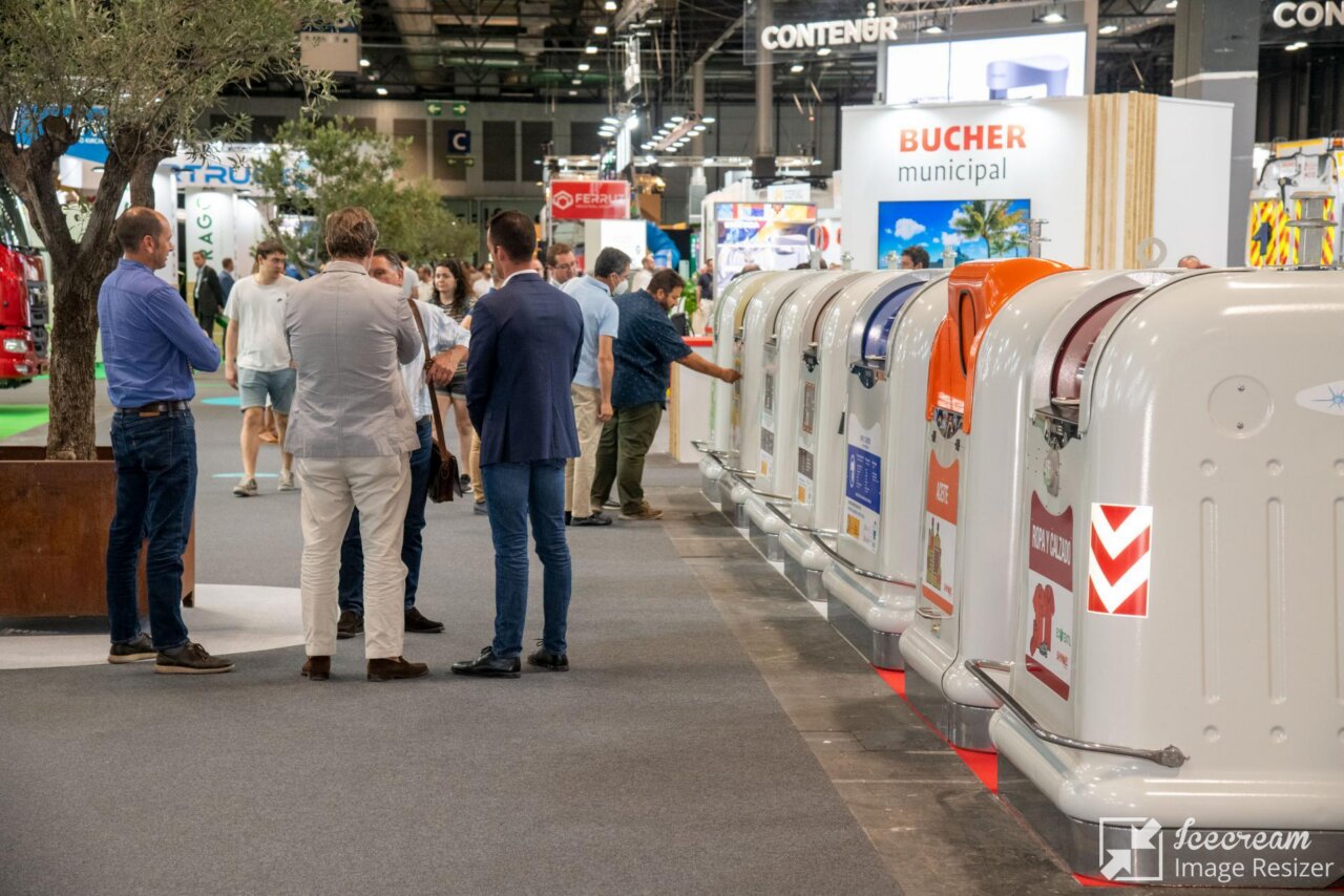 Group of professionals at an expo conversing near a row of large white curved municipal equipment or booths lined up on the right side of the hall.
