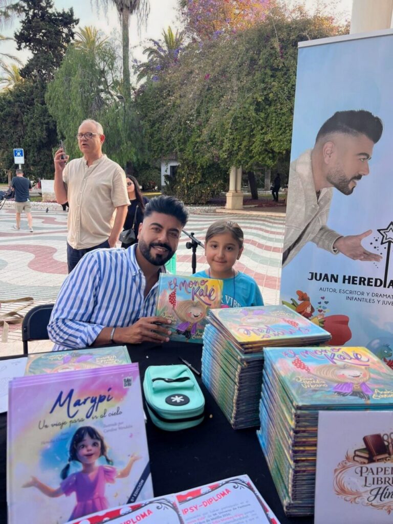 Author with a young girl at an outdoor book booth, smiling beside stacks of colorful children's books and a large poster on the right.