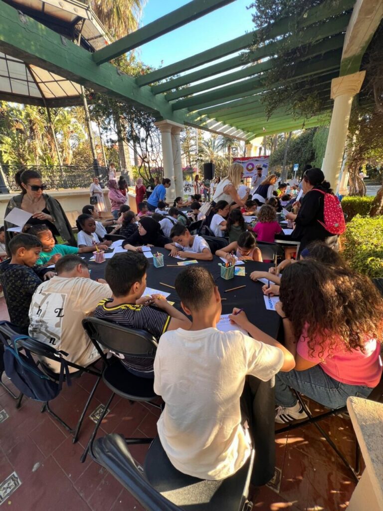 Group of children seated at an outdoor table drawing with pencils under a green pergola, with adults nearby supervising.