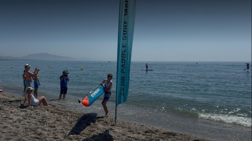 Girl on a pebbled beach carries a blue and orange inflatable buoy while a man films and others watch; a tall blue banner stands near the water.