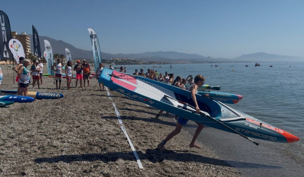 Group of people on a pebble beach carrying colorful windsurfing boards toward the water, with flags and spectators in the background.
