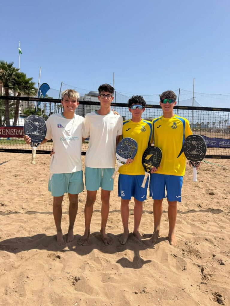 Four young players stand on a sandy beach tennis court, posing with paddles and smiling at the camera.