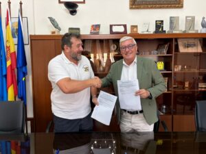 Dos hombres firmando un convenio en una oficina con banderas de fondo