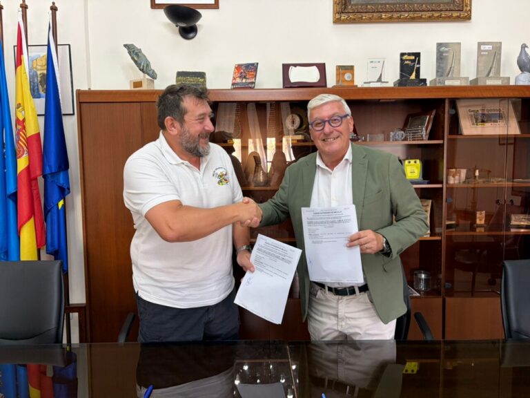 Dos hombres firmando un convenio en una oficina con banderas de fondo