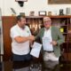 Dos hombres firmando un convenio en una oficina con banderas de fondo