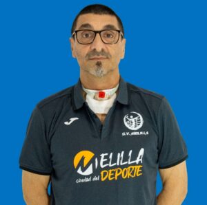 Portrait of a man with glasses wearing a dark polo featuring sports logos, with a visible tracheostomy tube on his neck against a solid blue background.