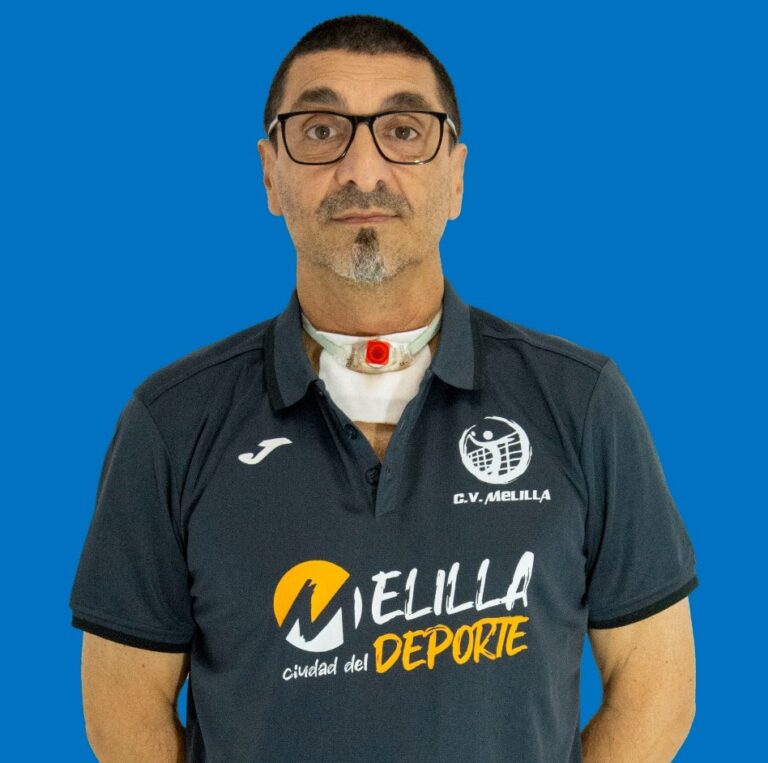 Portrait of a man with glasses wearing a dark polo featuring sports logos, with a visible tracheostomy tube on his neck against a solid blue background.