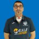 Portrait of a man with glasses wearing a dark polo featuring sports logos, with a visible tracheostomy tube on his neck against a solid blue background.