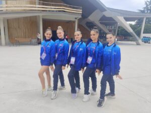 Five young female dancers in blue jackets posing outdoors on a concrete plaza.