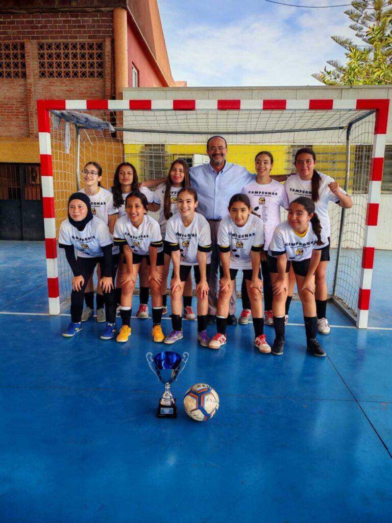 Girls' soccer team with coach posing in front of a goal on a blue court; trophy and ball in foreground.