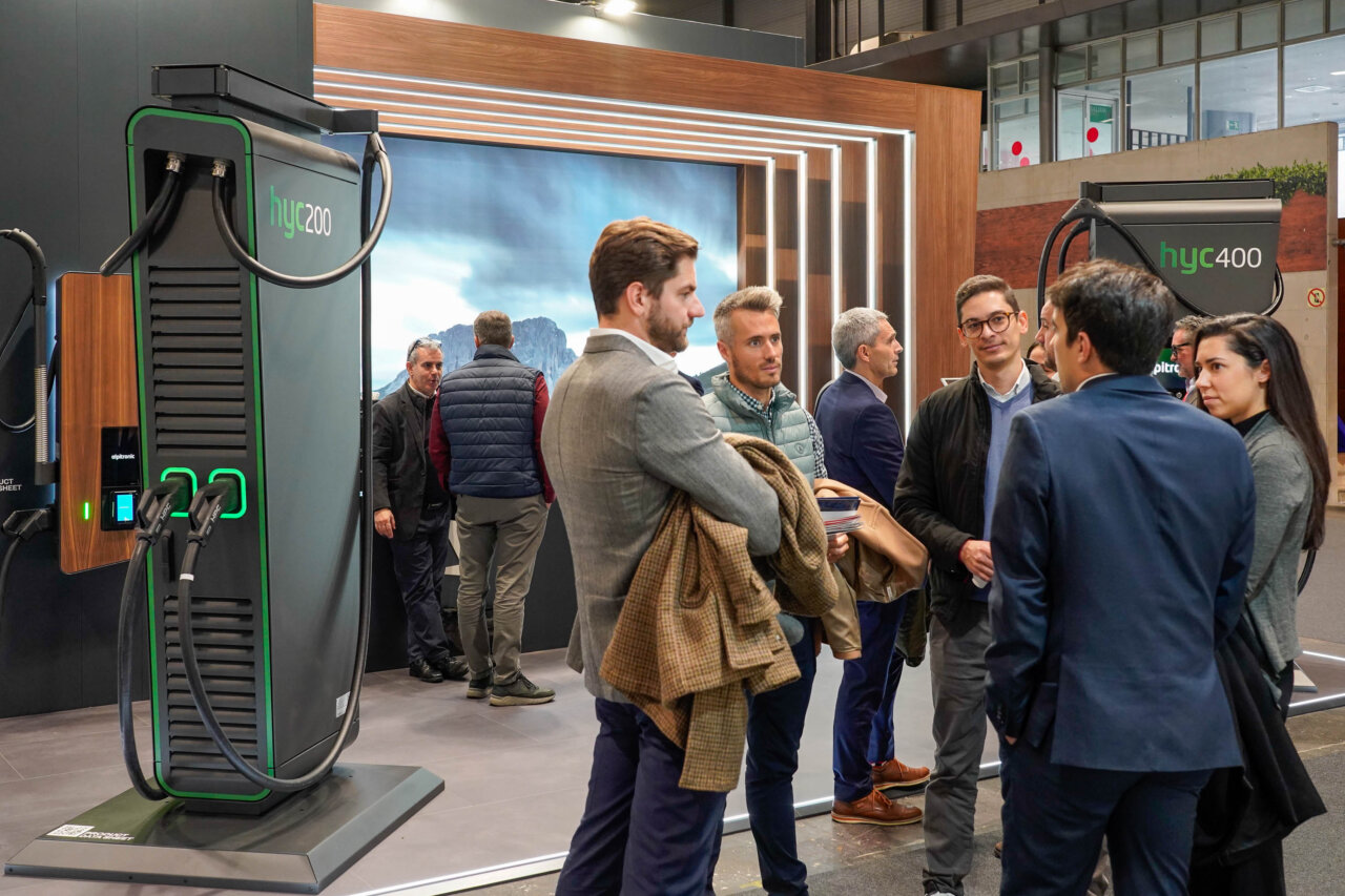 Group of professionals conversing at a technology expo, beside large green-and-black charging stations labeled hyc200 and hyc400.