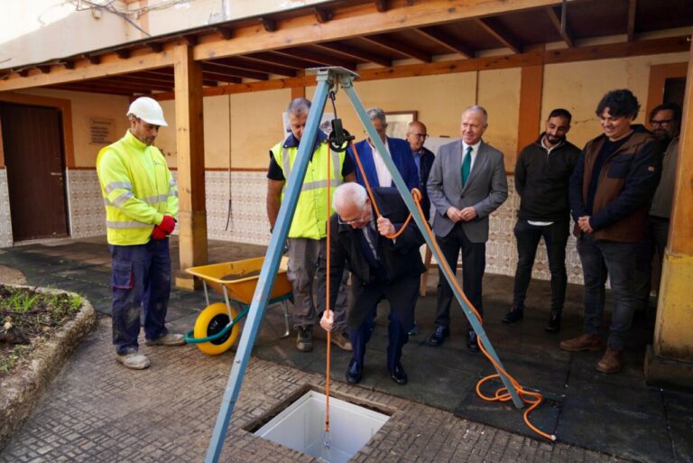 Group of workers and officials watching a man in a suit lowering a rope into an open manhole with a lifting tripod at a construction site during an inspection.