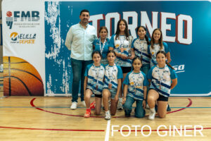 Group of young female basketball players in blue and white uniforms with medals, kneeling and standing with a coach in a gym, tournament banner in the background.