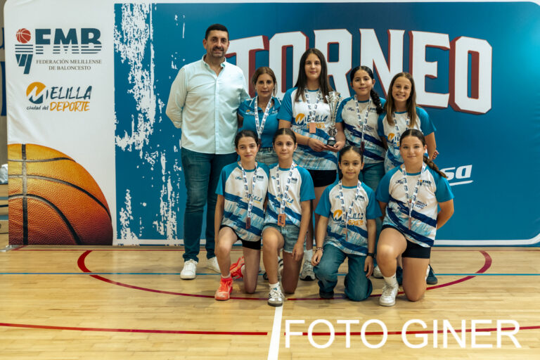 Group of young female basketball players in blue and white uniforms with medals, kneeling and standing with a coach in a gym, tournament banner in the background.